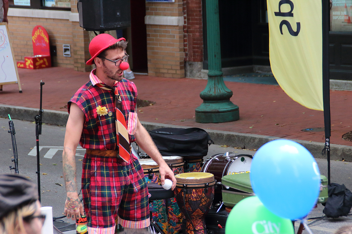 Street fair performer wearing red plaid suit with tie, hat, and red clown nose