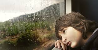 Child with sad expression staring at reflection in a rainy window