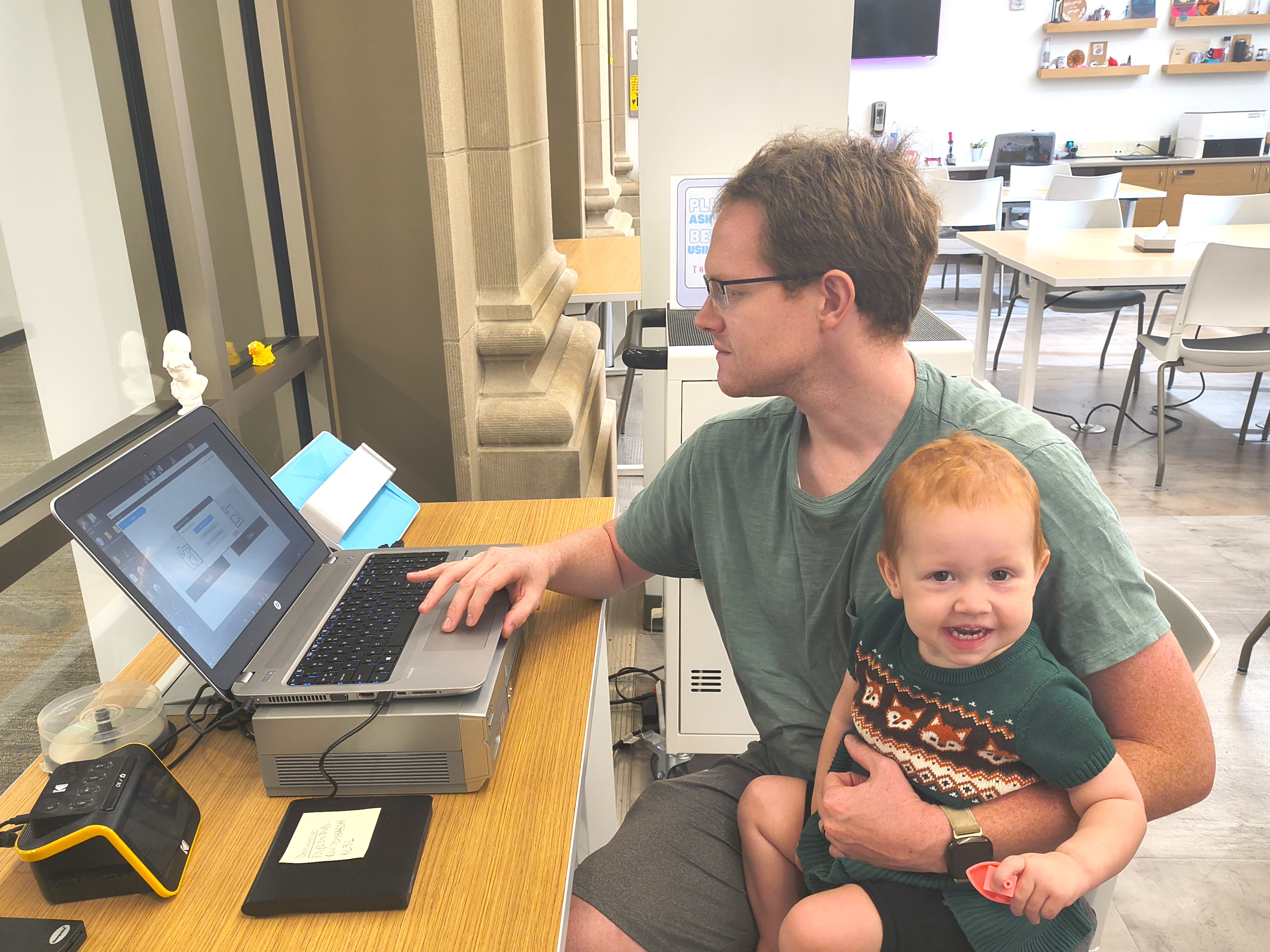 Photograph of an adult patron using the media converter in the IDEA Lab while holding a smiling baby
