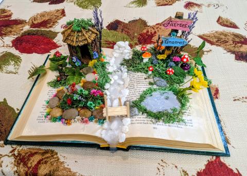 There is an open book decorated like a fairy garden with a hut, sign, moss, mushrooms and stones