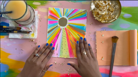 hands showing off a colorful neon sun painting with gold leaf, on a table with paints and craft supplies