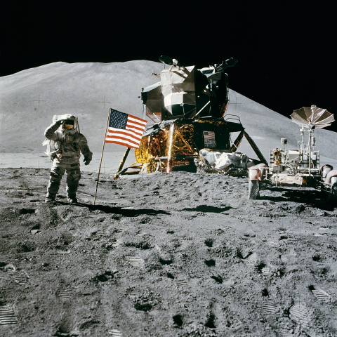 Astronaut saluting the camera next to an American flag in front of a space craft and a lunar rover on the moon