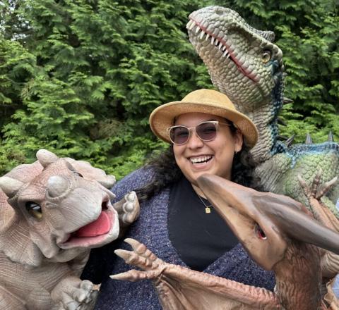 Woman surrounded by three dinosaurs with background of greenery