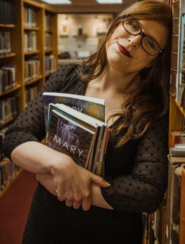 Sara Henning stands next to a book shelf wearing a black dress, holding a stack of books and smiling