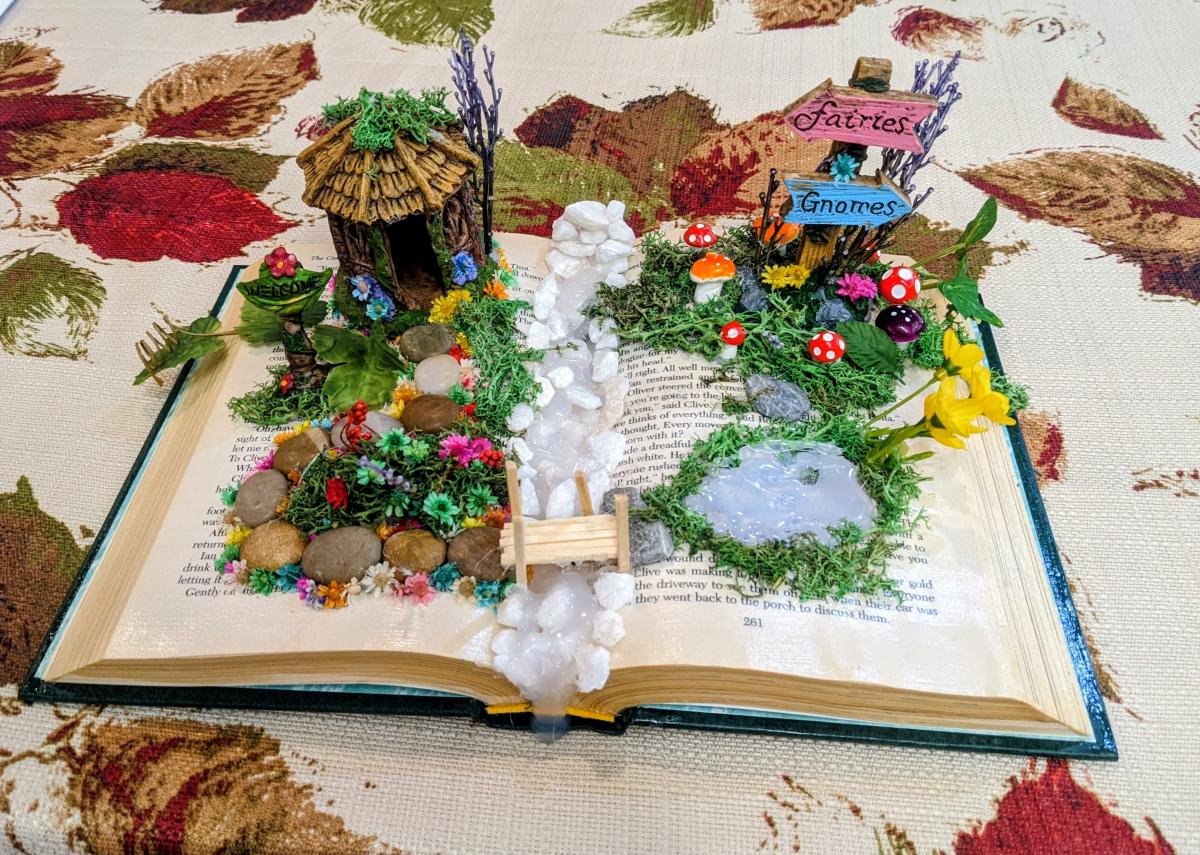 There is an open book decorated like a fairy garden with a hut, sign, moss, mushrooms and stones