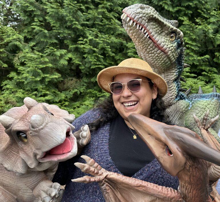 Woman surrounded by three dinosaurs with background of greenery